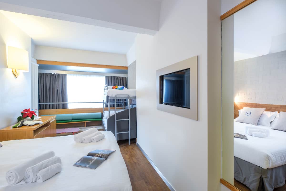 A hotel room with a bunk bed, a desk with a lamp, a bed with towels, and a television on the wall.