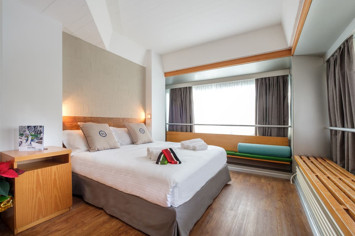 A modern hotel room with a large bed, a wooden nightstand, a bench, and a window with curtains.