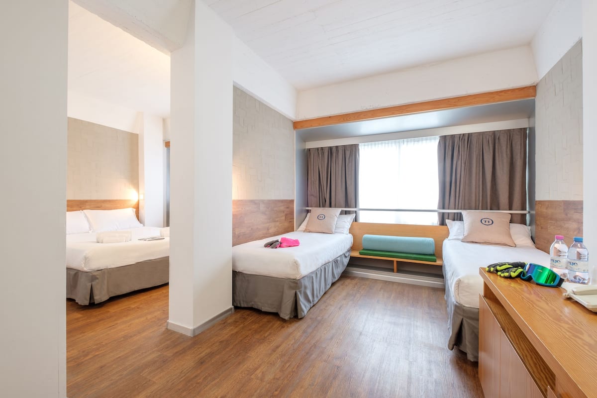 A hotel room with two beds, a wooden desk, a window with curtains, and a view of the outside.