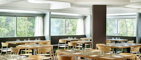 A dining area with multiple tables and chairs, large windows with curtains, and a high ceiling with recessed lighting.