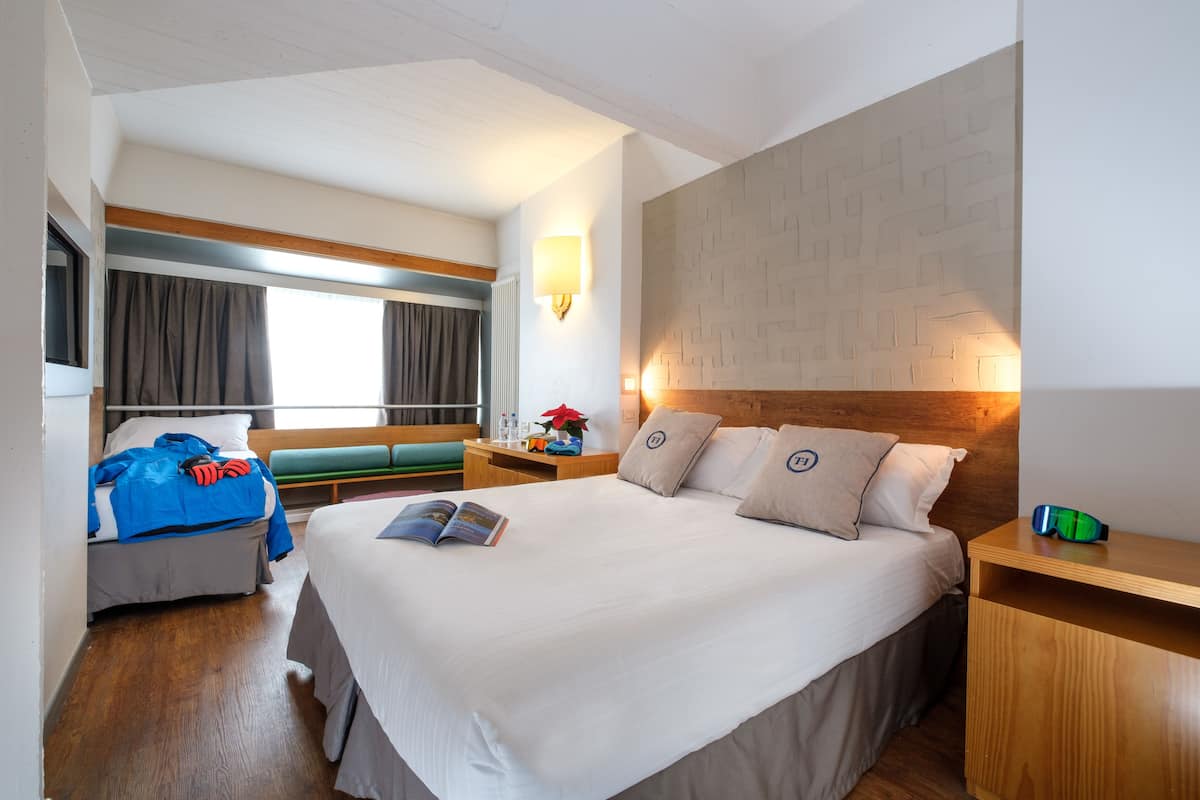 A hotel room with a large bed, a bedside table, a nightstand, a window with curtains, and a wall with a geometric pattern.