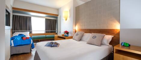 A hotel room with a large bed, a bedside table, a nightstand, a window with curtains, and a wall with a geometric pattern.