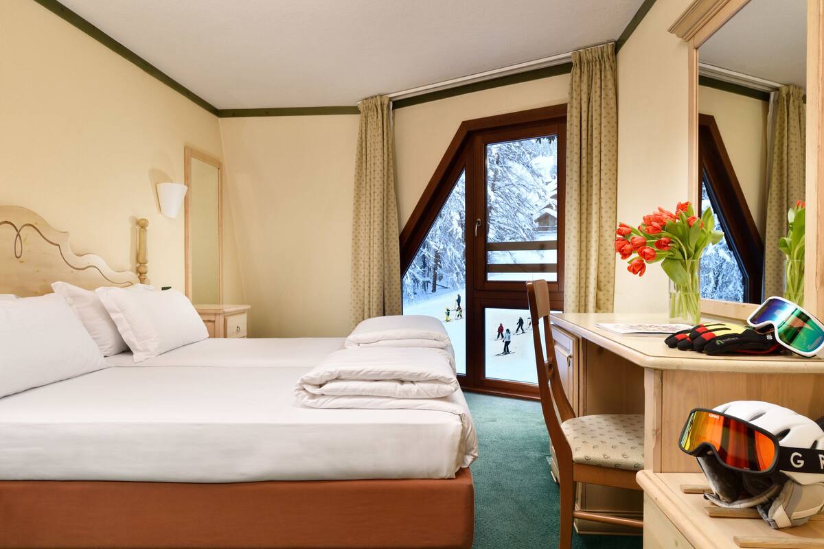 Una camera d'albergo con un letto grande, una scrivania con occhiali da sci e vista su un paesaggio innevato.