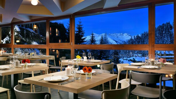 Ein Restaurant mit gedeckten Tischen und großen Fenstern, durch die sich verschneite Berge zeigen.