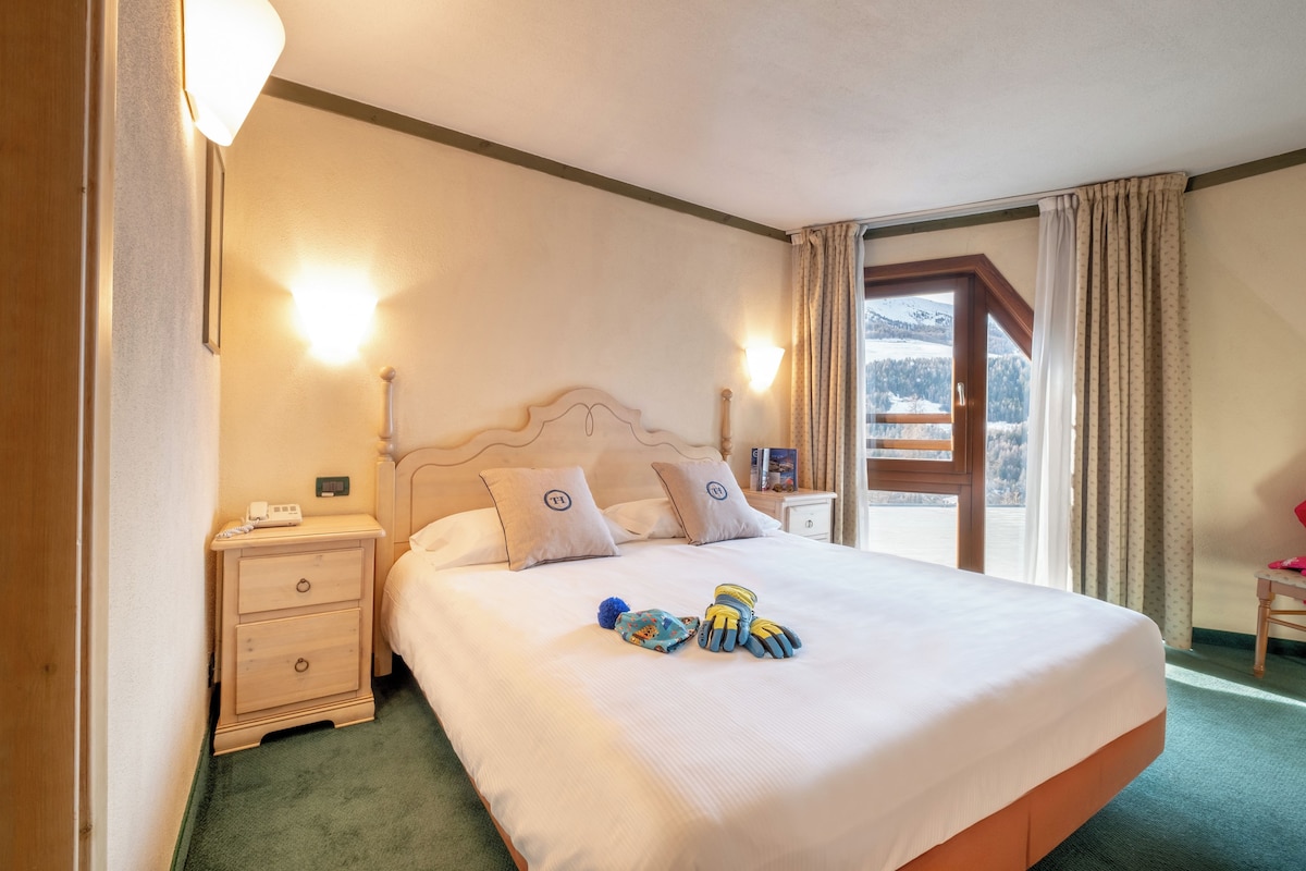 A hotel room with a large bed, two bedside tables, a nightstand, and a view of a mountain landscape through a window.