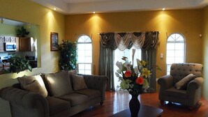 A living room with a sofa, armchair, and a vase of flowers. There is a window with curtains and a plant on the left. - Faith's Villa of Tobago (Scarborough)