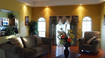 Sala de estar com sofá, poltrona e um vaso de flores. Há uma janela com cortinas e uma planta à esquerda.
