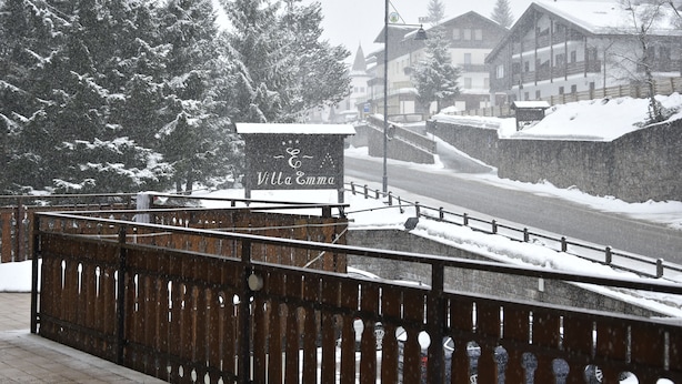 A snow-covered outdoor area with a wooden fence, a sign for "Villa Emma," and buildings in the background. - IHR Hotel Villa Emma (Canazei)