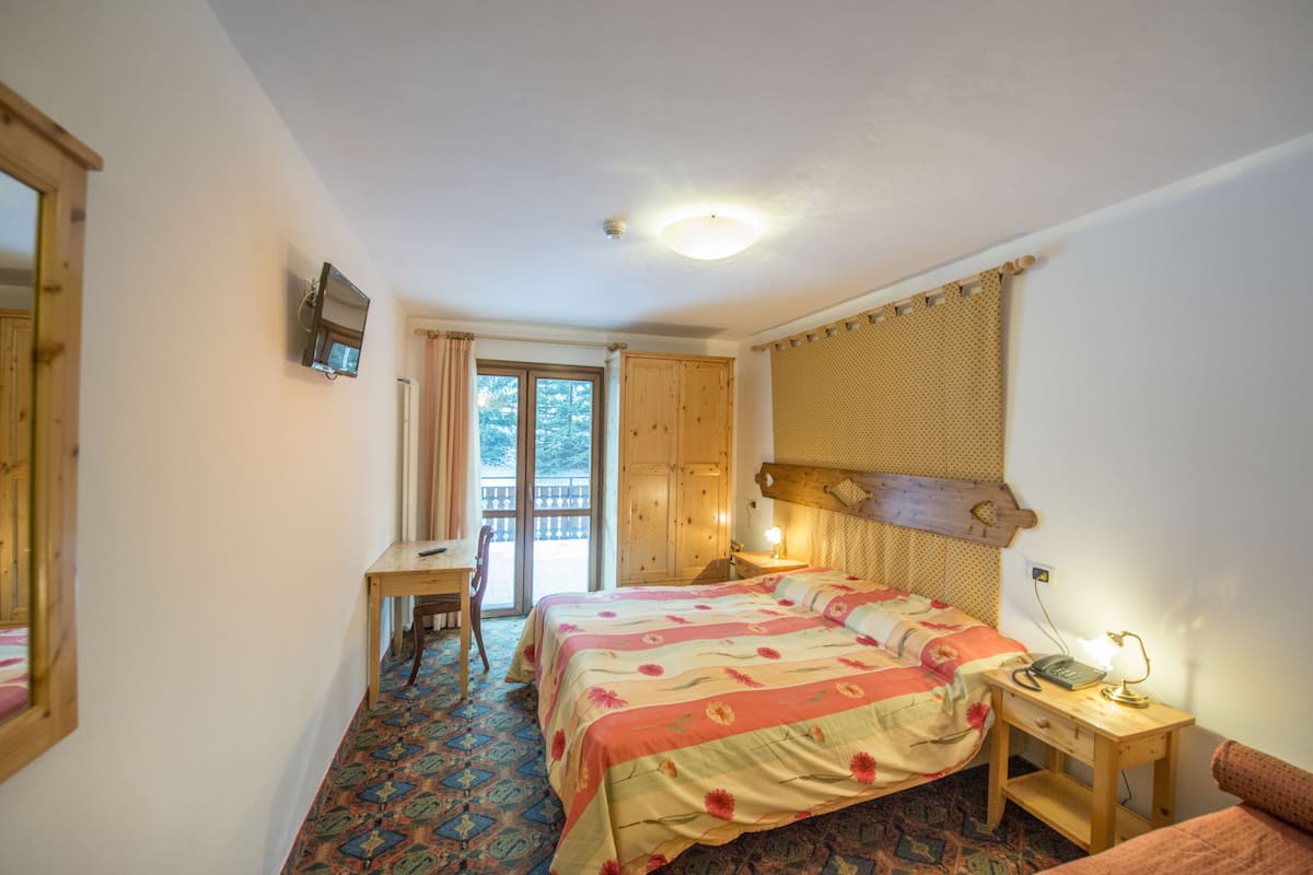 A bedroom with a bed, a desk, a chair, a bedside table with a lamp, and a window with curtains.