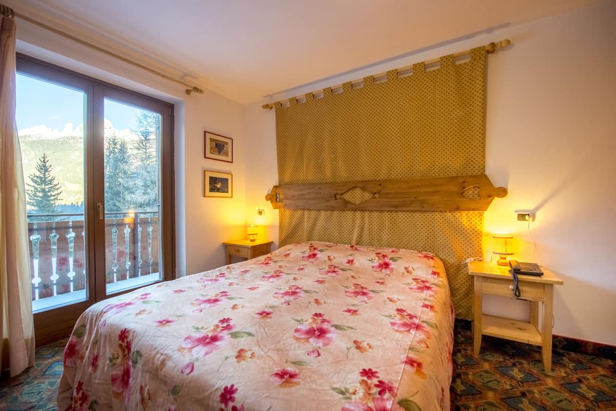 A bedroom with a floral bedspread, a wooden headboard, a balcony with a view of trees, and two bedside lamps.