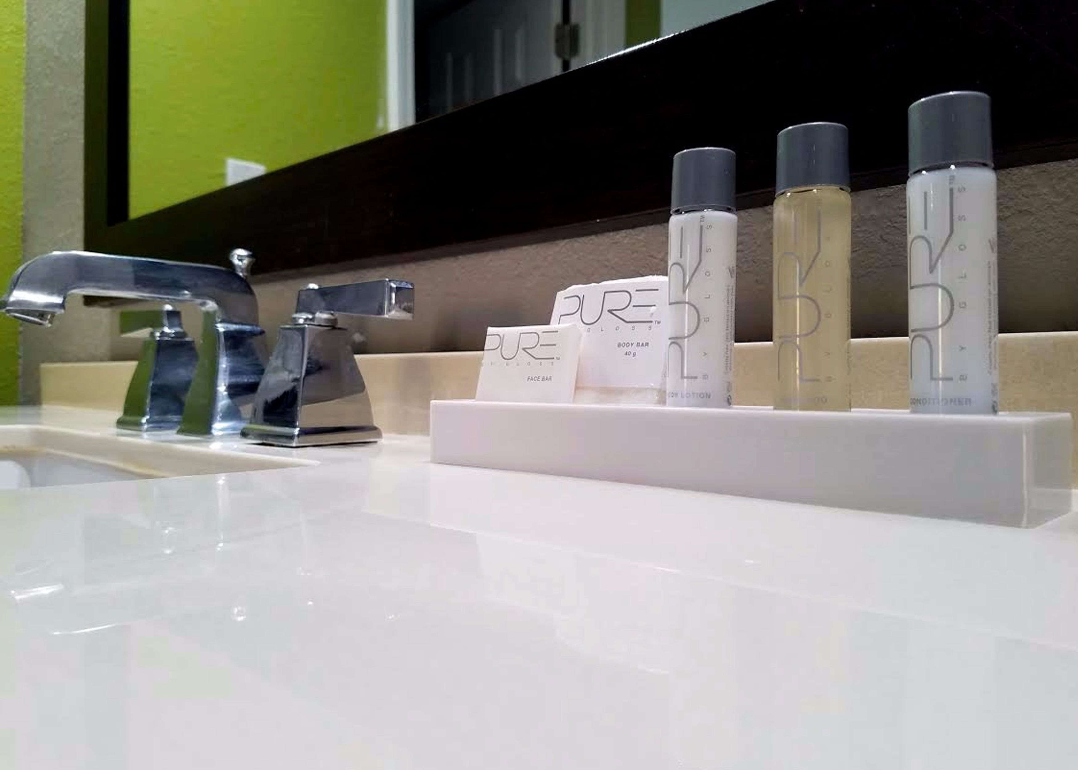 A bathroom sink with a chrome faucet and a mirror above it. On the sink, there are several bottles of 'PURE' body care products, including a body bar, lotion, and conditioner.