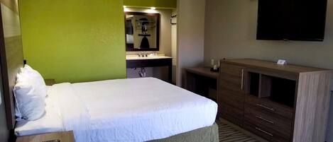 A hotel room with a bed, a desk, a dresser, a mirror, a TV, and a bathroom with a sink and mirror.