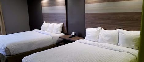 A hotel room with two beds, each with white bedding and pillows, a wooden headboard, and a nightstand with a lamp.