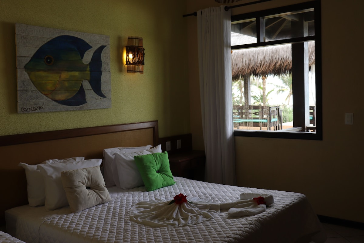 Habitación de hotel con cama, almohadas, un cuadro, ventana con vistas y lámpara fijada a la pared.
