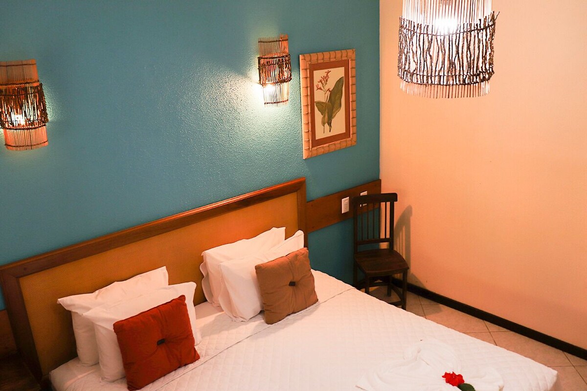 A neatly made bed with white linens and a single red rose, a wooden headboard, and a chair against a teal wall with framed botanical prints.