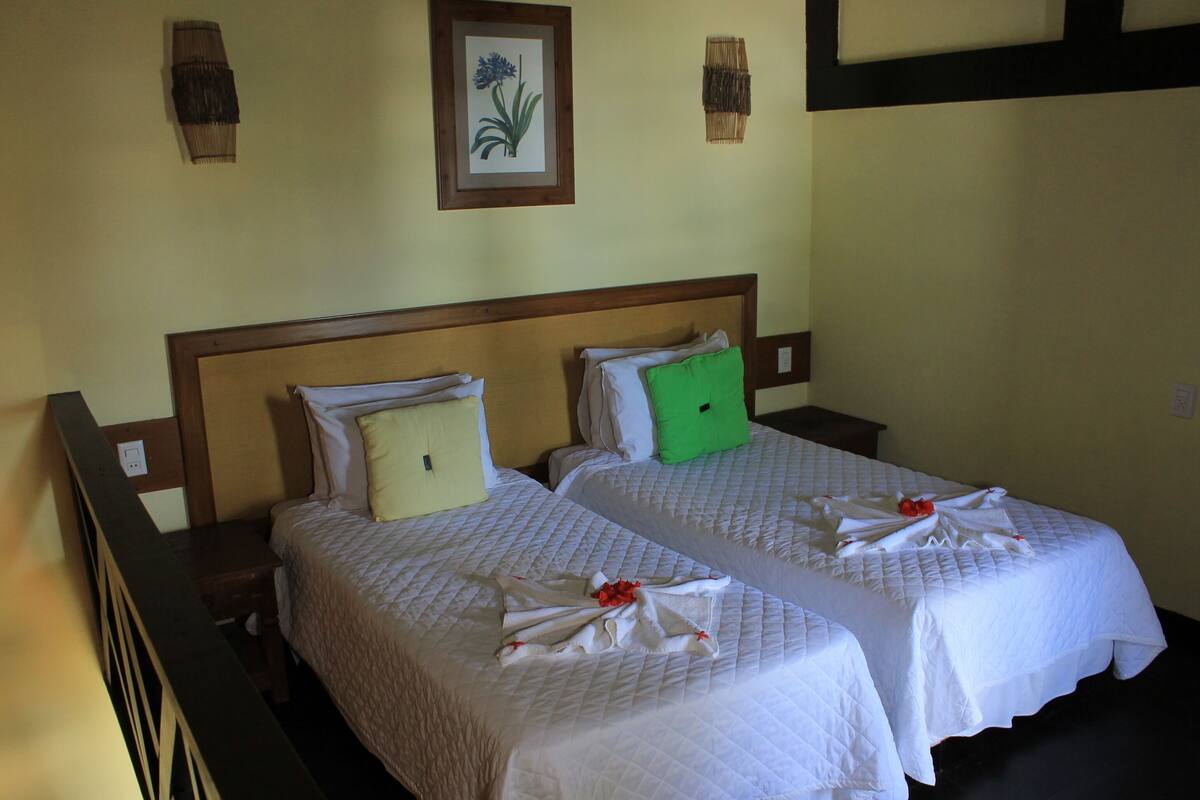 Ein Hotelzimmer mit zwei Betten, jeweils mit ordentlich bezogener Bettdecke und Kissen. Darüber an der Wand hängt ein gerahmtes Pflanzenbild.