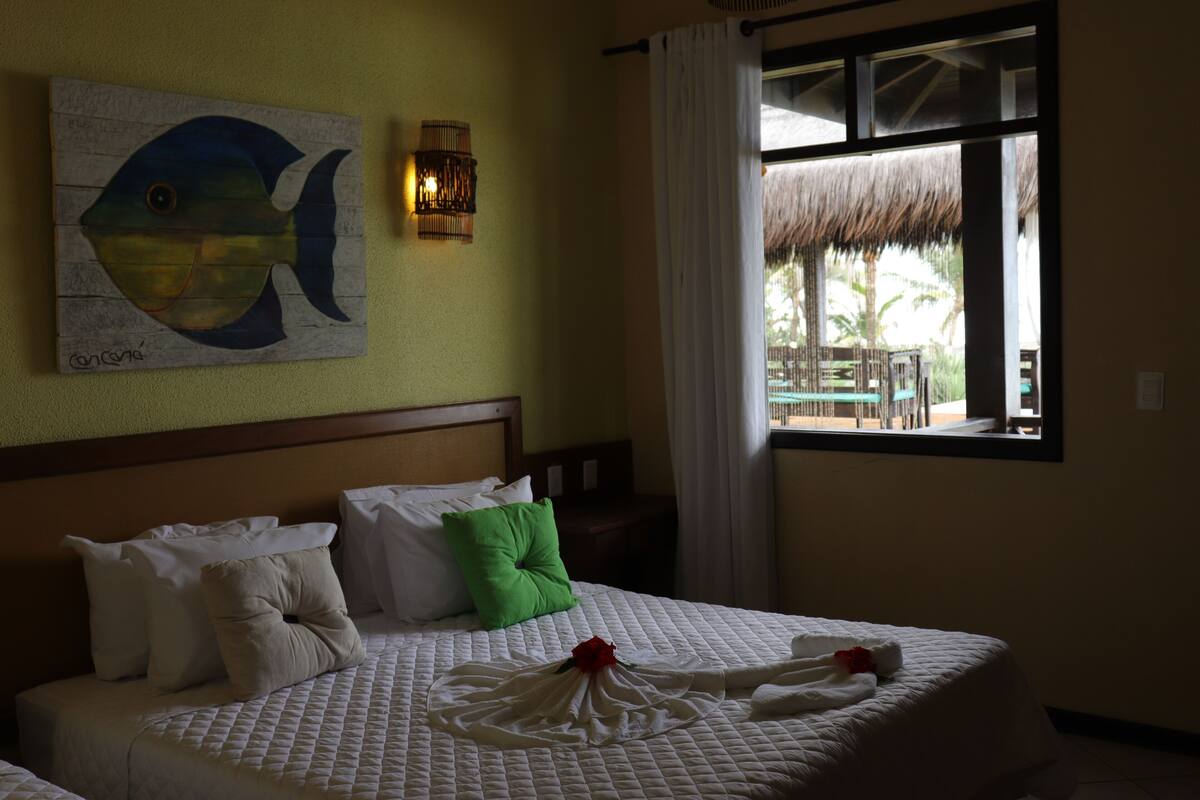 Ein Hotelzimmer mit einem Bett, Kissen, einem Gemälde, einem Fenster mit Ausblick und einer Wandleuchte.