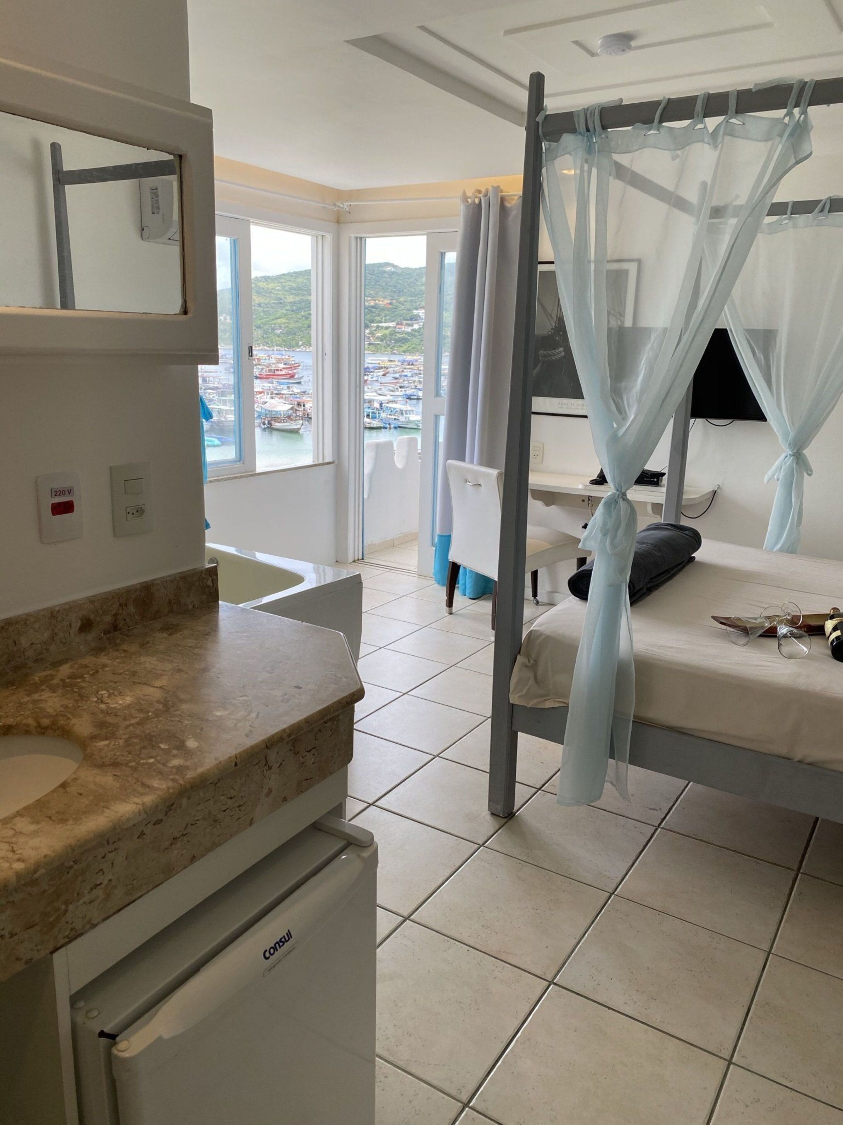 A bedroom with a bed, a bedside table, a small bedside lamp, a window with a view of boats, and a bathroom with a sink and mirror.
