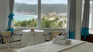 Une chambre d’hôtel avec un lit, une petite table ornée d’un coquillage, deux chaises et une vue sur une plage et des collines.