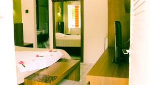 A hotel room with a bed, a bench, a mirror, a TV, and a bathroom visible through an open door.