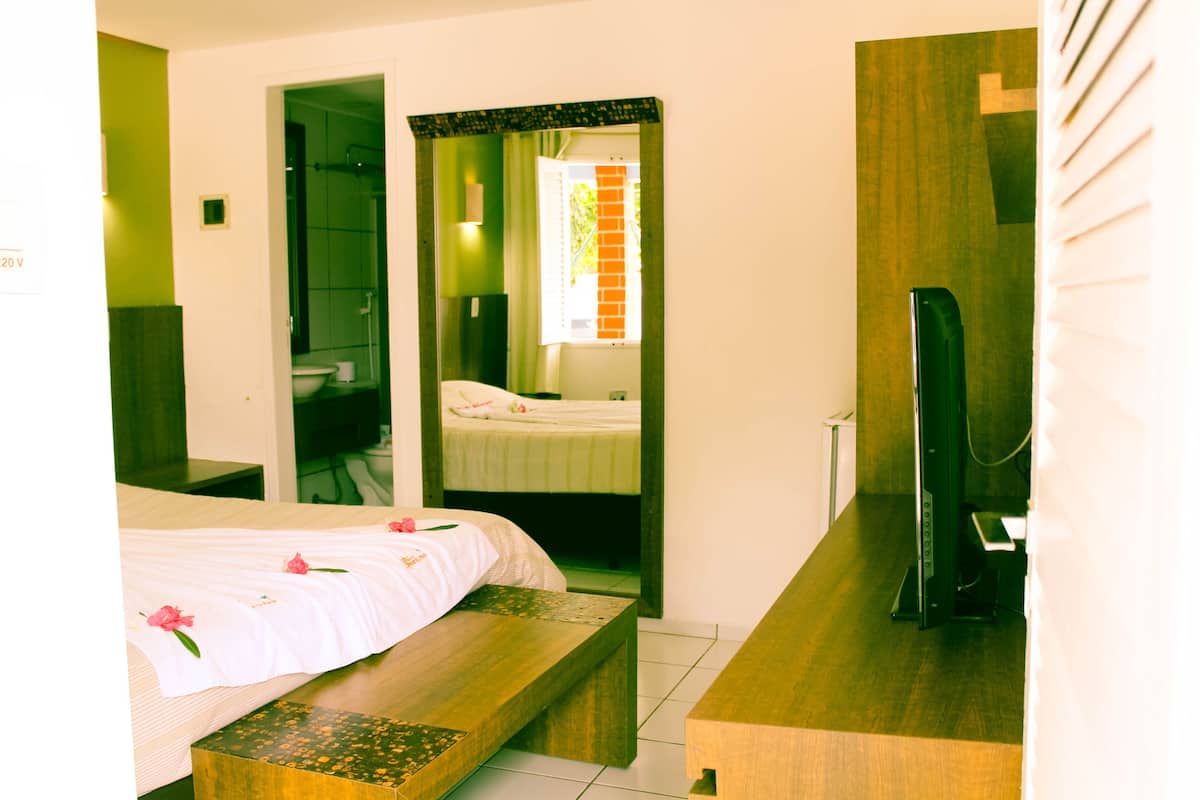 Ein Hotelzimmer mit Bett, Sitzbank, Spiegel, Fernseher und einem durchgegangenen Tür sichtbaren Badezimmer.