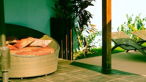 A wicker sofa with a cushion, a wicker chair, and a potted plant in a room with a tiled floor.