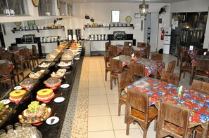 Un comedor tipo bufé con varias mesas y sillas, una zona de cocina con electrodomésticos y estanterías, y un suelo de baldosas.