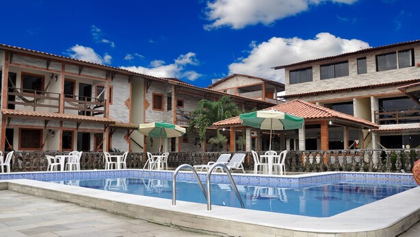 A swimming pool with lounge chairs and umbrellas, surrounded by multi-story buildings with balconies and windows. - Pousada Porto Familly (Ipojuca)