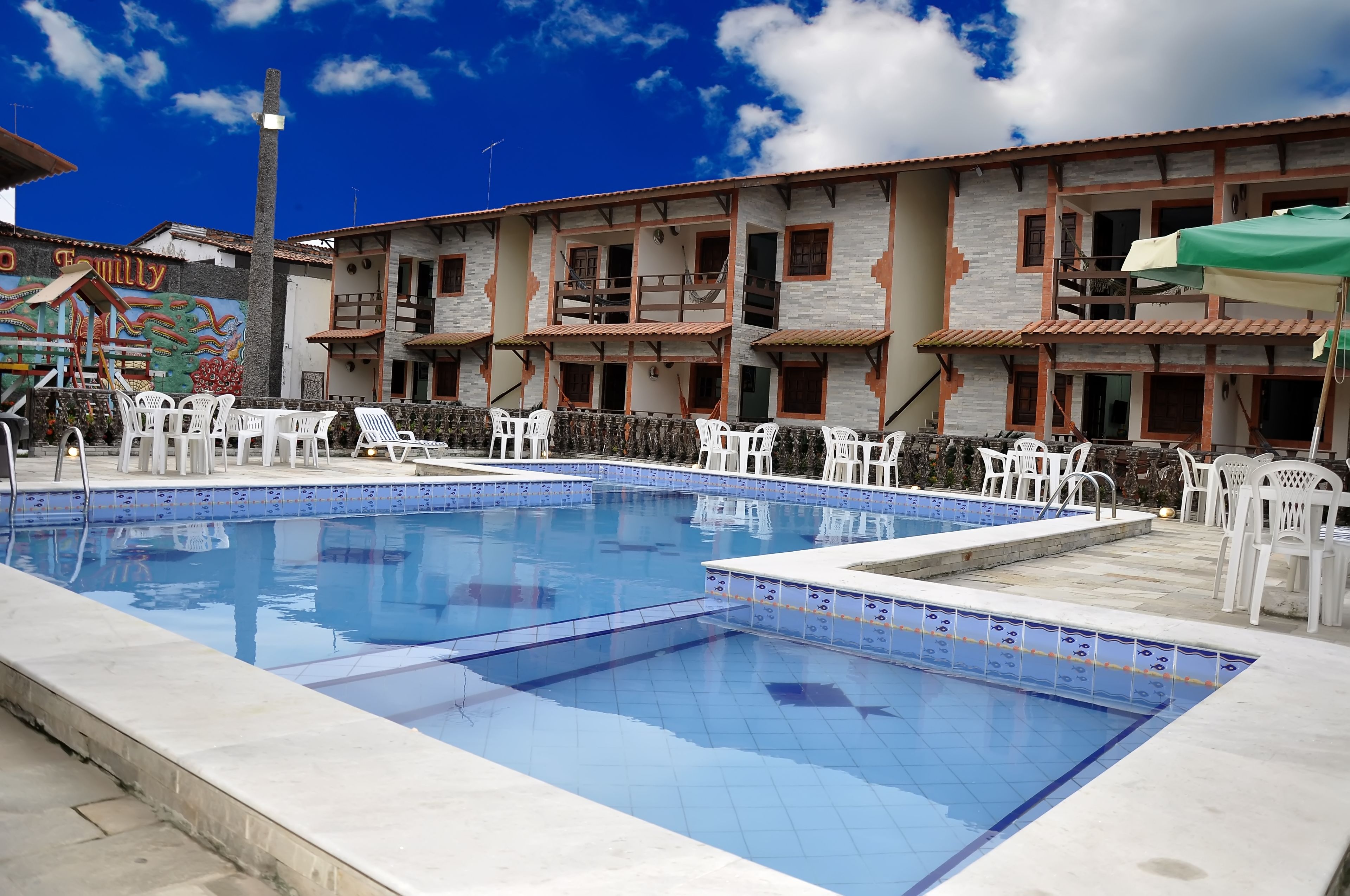 A swimming pool with white chairs, a building with a balcony, and a mural.