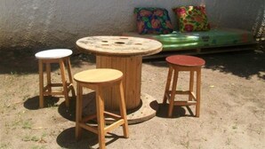 A round wooden table with four stools, a green sofa with colorful cushions, and a concrete wall in the background. - Hostel Guerreiro (Maceió)
