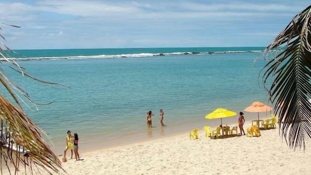 A beach with clear turquoise water, a sandy shore, and a few people enjoying the water. There are yellow beach chairs and umbrellas set up for sunbathing. - Hostel Guerreiro (Maceió)