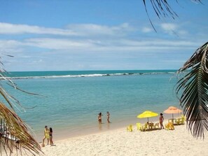 Una playa con agua turquesa cristalina, una orilla de arena y algunas personas disfrutando del agua. Hay sillas de playa y sombrillas amarillas dispuestas para tomar el sol.