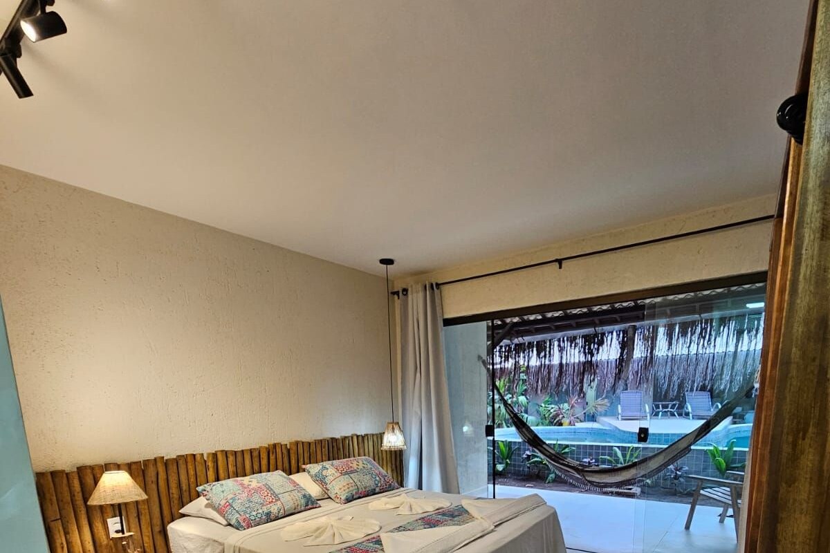 A bedroom with a large bed, a nightstand, a lamp, and a view of a pool and hammock outside.