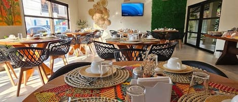 A dining area with round tables set with plates, cutlery, and woven placemats. There are hanging pendant lights, a television on the wall, and a green wall feature.