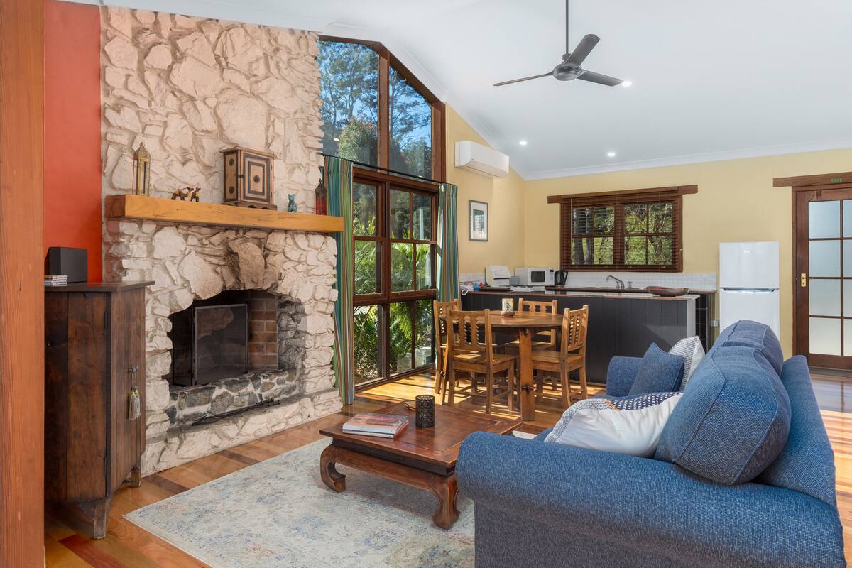A living room with a stone fireplace, a blue sofa, a wooden coffee table, and a dining area with wooden chairs and a table.
