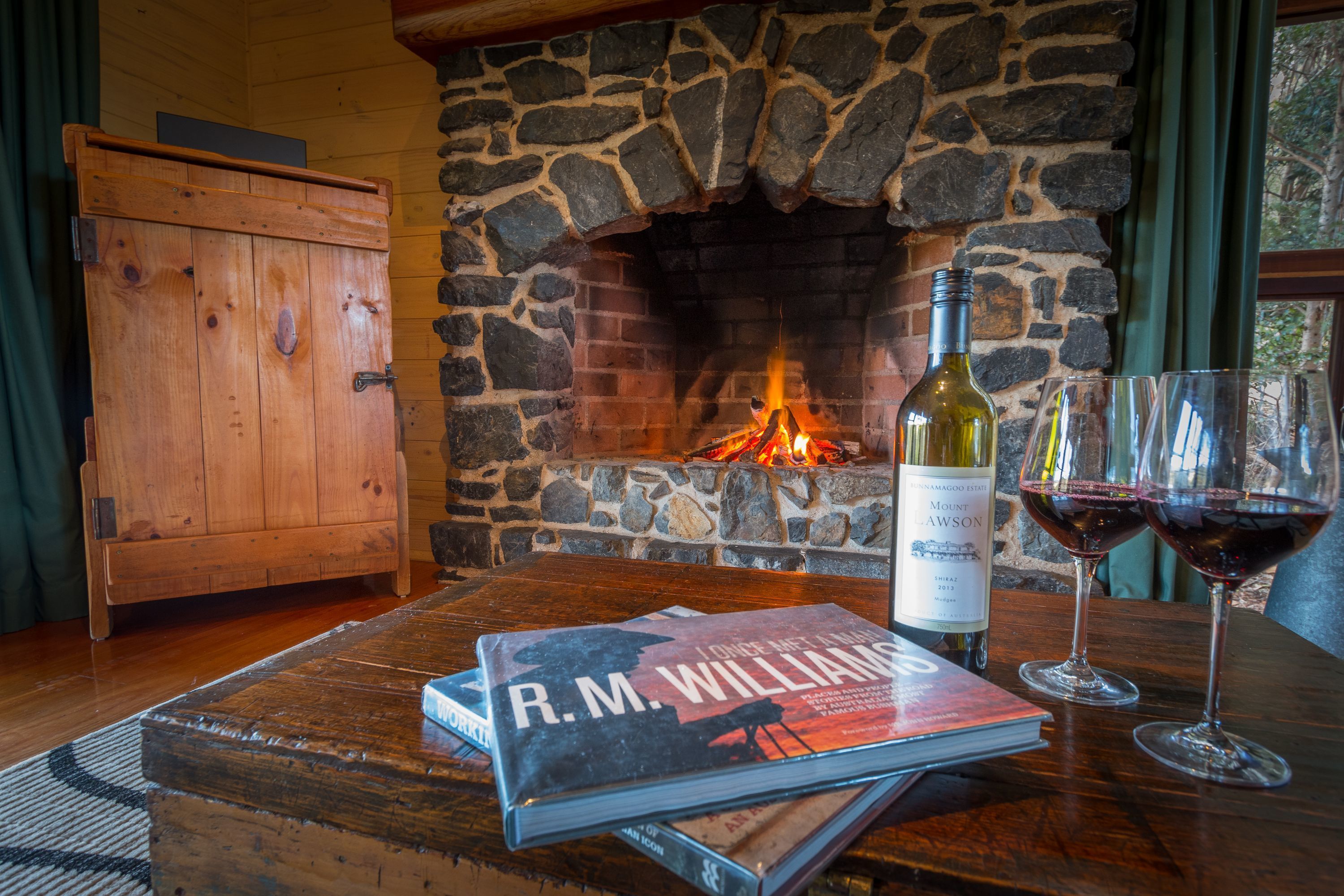 A cozy room with a stone fireplace, a wooden table with a book and wine glasses, and a wooden cabinet.