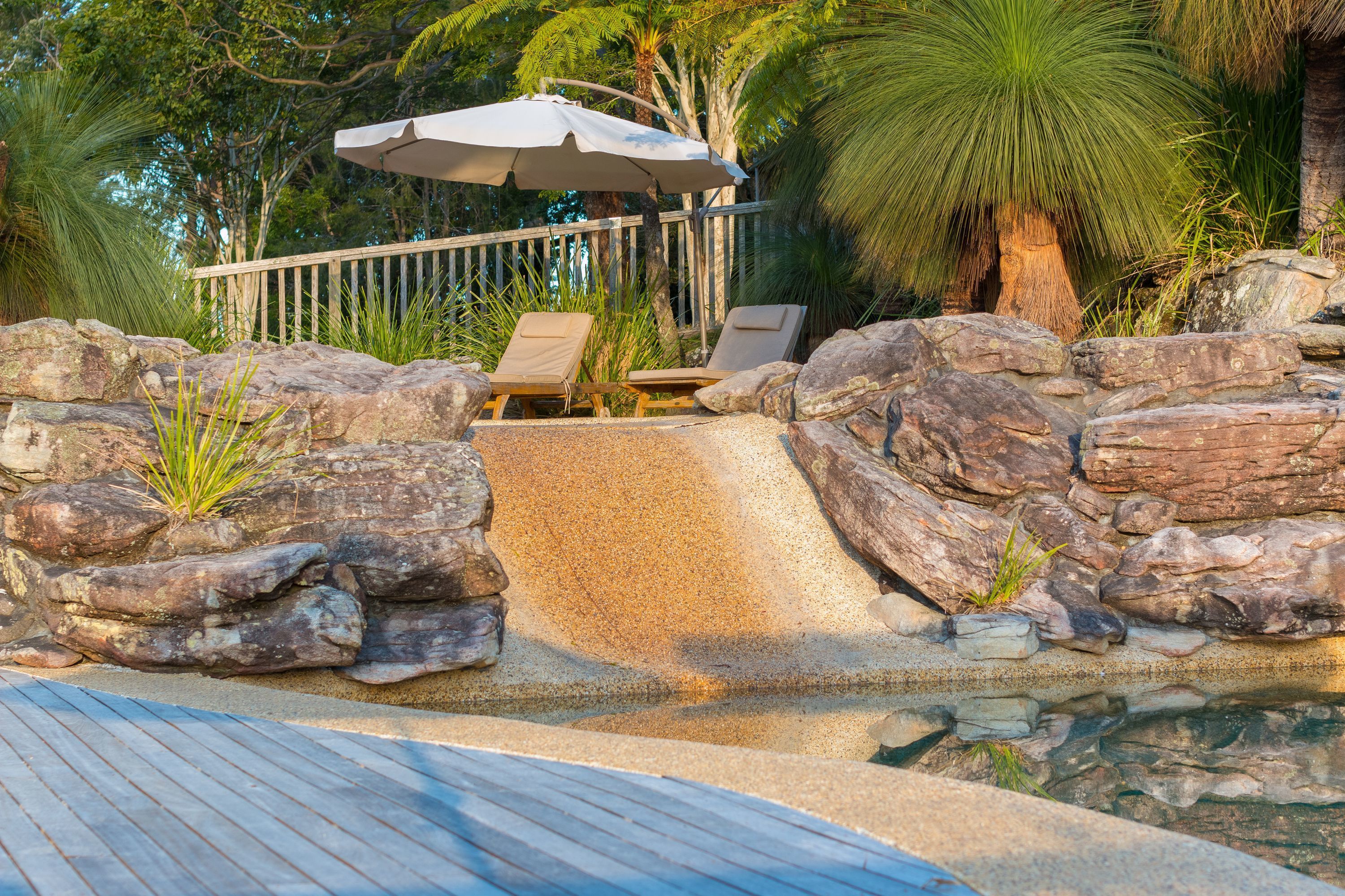 A pool area with lounge chairs and an umbrella, surrounded by rocks and plants.