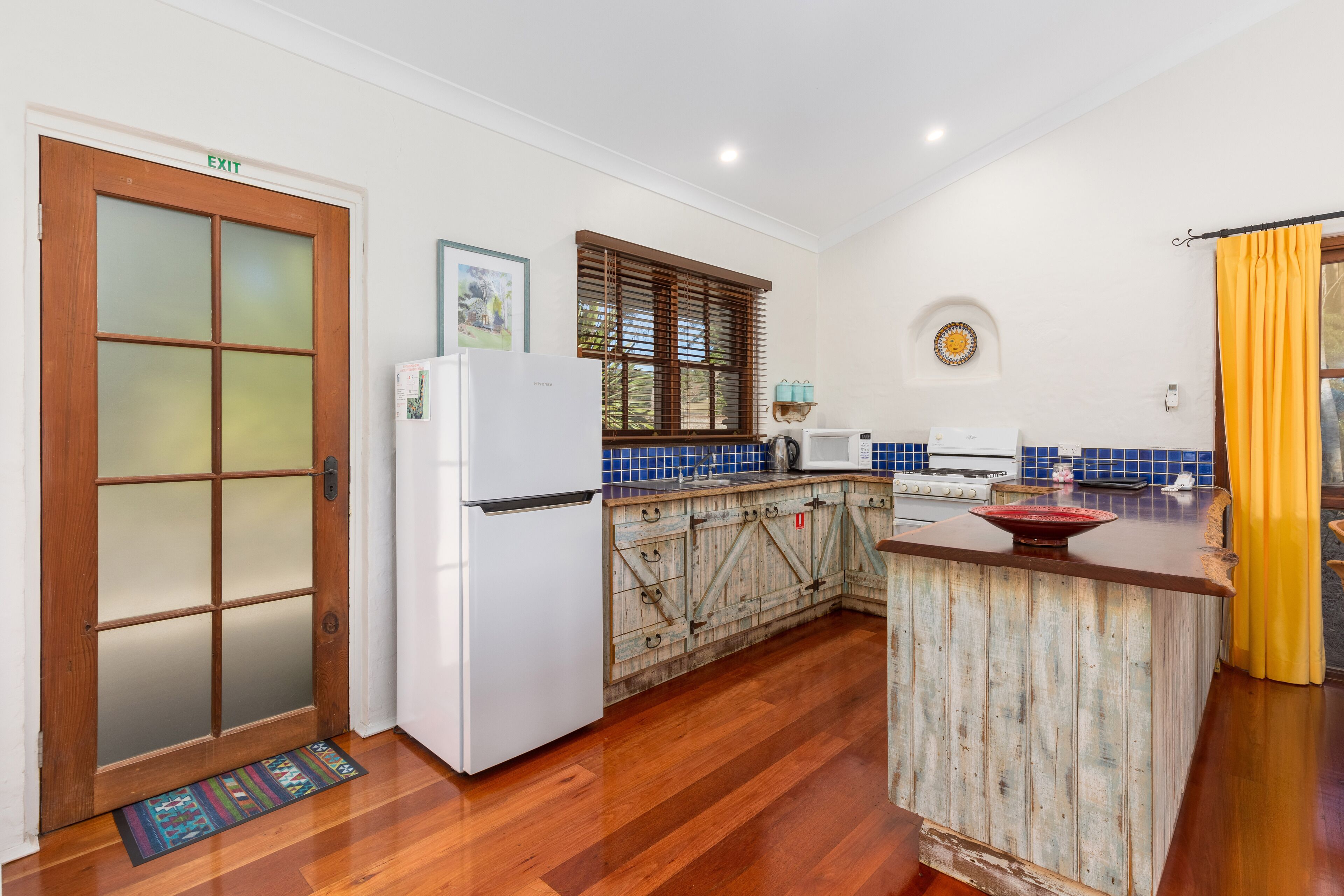 A kitchen with a wooden door marked 'EXIT', a white refrigerator, a blue tiled backsplash, a window with wooden blinds, and a yellow curtain.