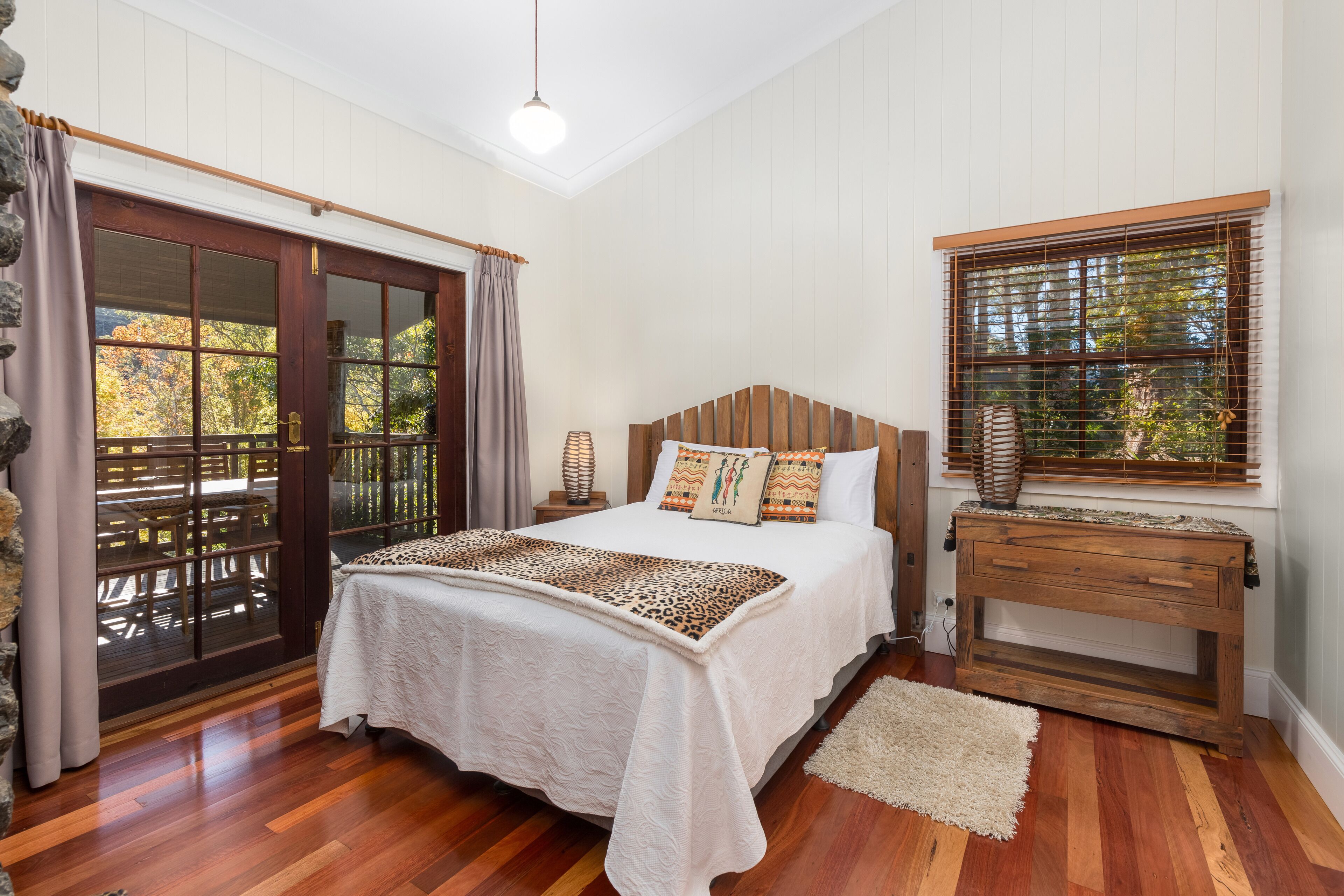 A bedroom with a wooden bed, a nightstand, a window with blinds, and a view of the outdoors.