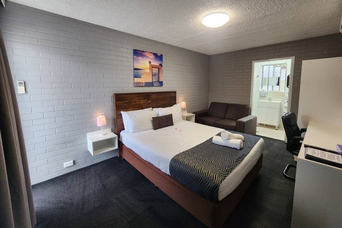 Habitación de hotel con cama, mesita de noche, escritorio, silla y baño visible a través de una puerta abierta.