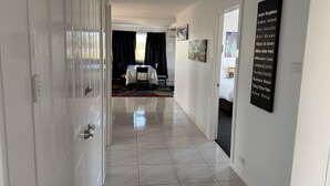 A hallway with a dining area in the background, a bed with a white headboard, and a wall with a list of names. - Crest Motor Inn (Queanbeyan)