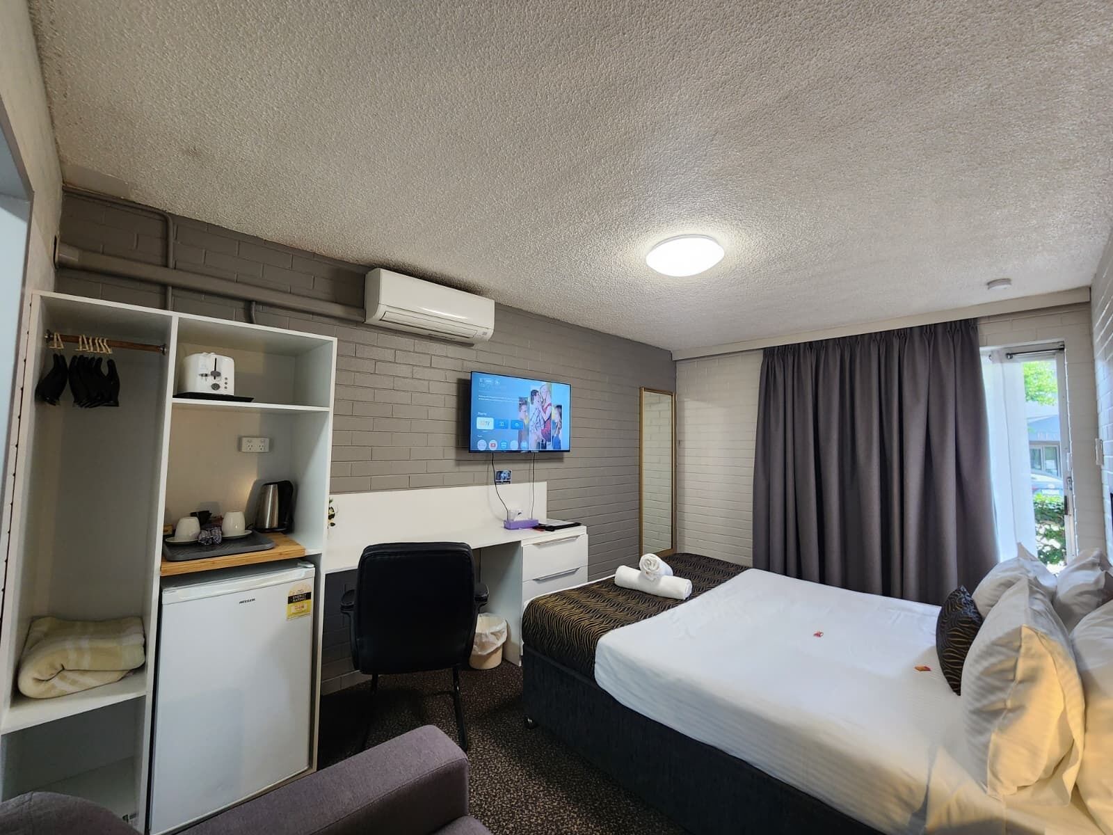 Une chambre d’hôtel comprenant un lit, un bureau avec un ordinateur, une télévision, une armoire et une petite kitchenette.