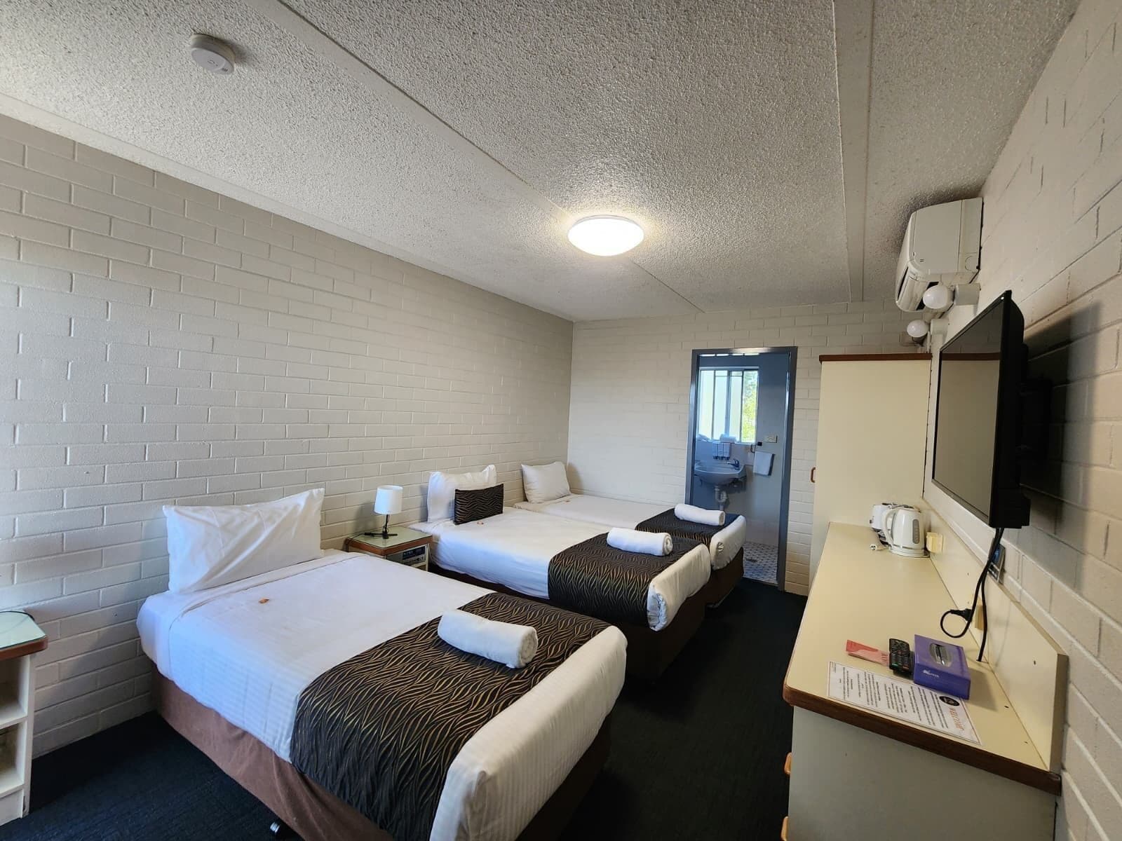 A hotel room with two beds, a desk with a computer and a telephone, a television, and a bathroom visible through a door.