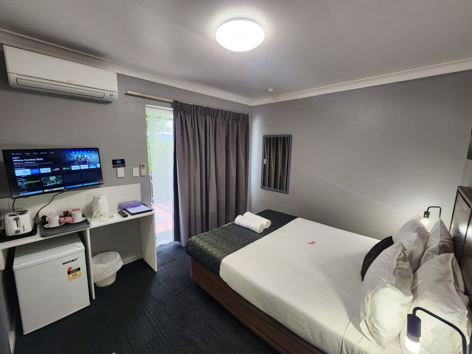 Quarto de hotel com cama, escrivaninha com televisão, máquina de café e lixeira.