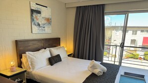A hotel room with a bed, a headboard, a nightstand with a lamp, a window with curtains, and a view of the outside. - Crest Motor Inn (Queanbeyan)