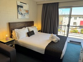 Habitación de hotel con cama, cabecera, mesita de noche con lámpara, ventana con cortinas y vista al exterior.