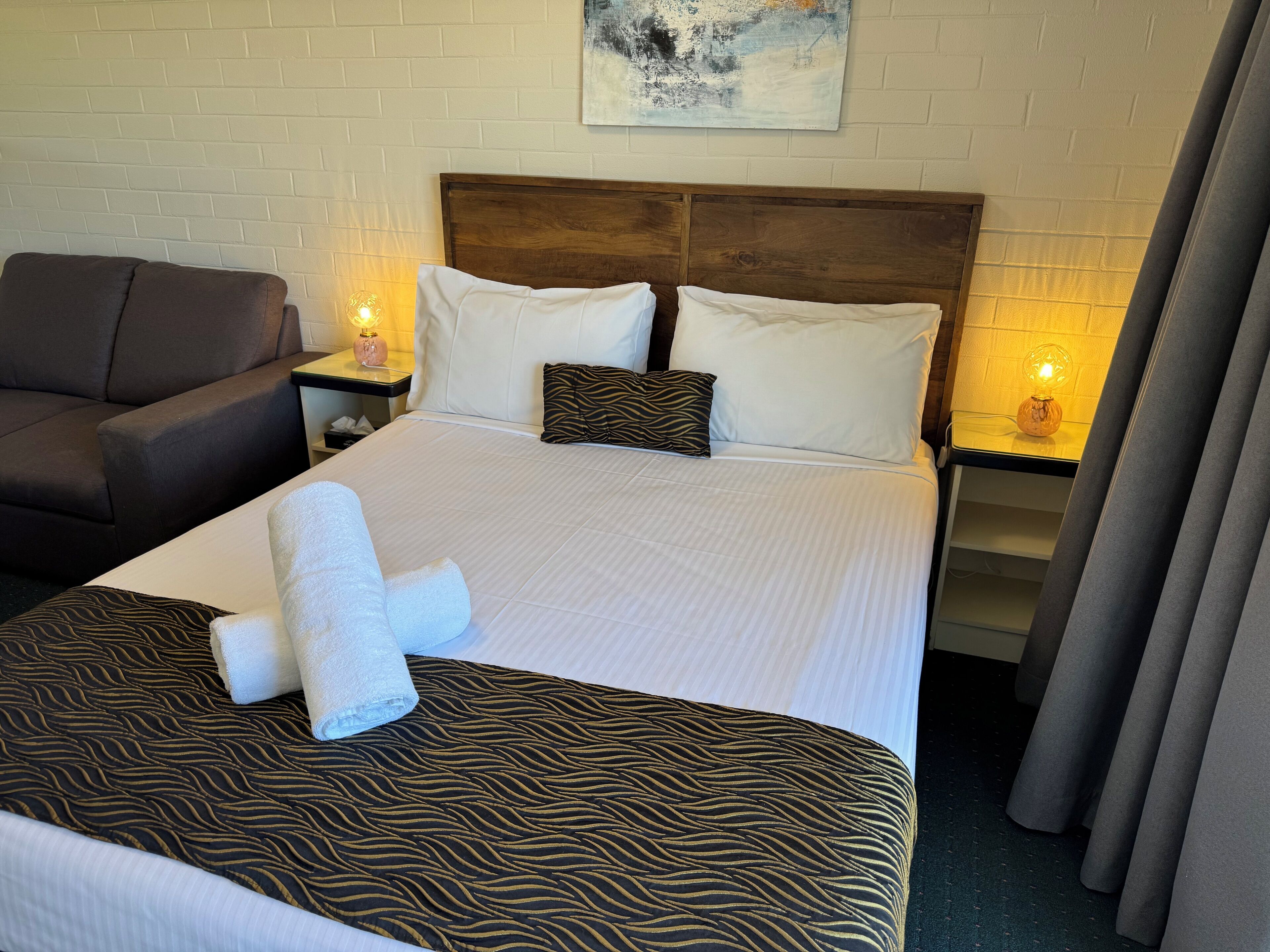 Et hotelværelse med en seng, en stol, et natbord med lampe og et billede på væggen.