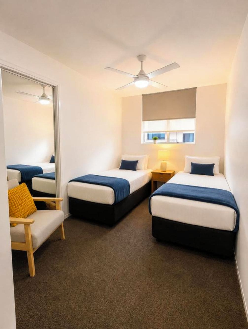 Une chambre d’hôtel avec deux lits simples, un ventilateur de plafond, une petite table avec une lampe et une fenêtre donnant sur un paysage agréable.
