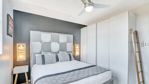 A modern bedroom with a tufted headboard, a standing bedside table, a wardrobe, and a ceiling fan. - Tradewinds Apartments (Coffs Harbour)