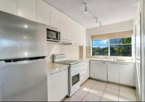 A modern kitchen with white cabinetry, a stainless steel refrigerator, a microwave, a stove, and a dishwasher. There is a window providing a view of greenery outside. - Tradewinds Apartments (Coffs Harbour)